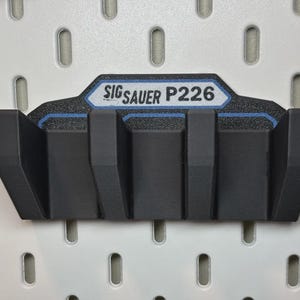 May include: A black, 3D-printed wall mount for a SIG Sauer P226 firearm. The mount features the text "SIG Sauer P226" in white on a blue and black background. The mount is designed to hold the firearm securely.