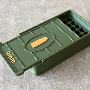 Może przedstawiać: Zielone pudełko do przechowywania amunicji 9 mm z klapką na zawiasach. Pokrywa ma owalny kształt z beżową grafiką pocisku. Tekst "9mm" jest wytłoczony złotem z przodu pudełka.