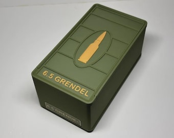 Può includere: Scatola rettangolare verde oliva con grafica a proiettile dorata. Il testo "6.5 Grendel" è stampato in oro sulla parte superiore e anteriore. La scatola è su una superficie bianca.