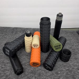 May include: An assortment of cylindrical suppressors in various colors, including black, orange, and green. Some have text labels such as "YHM R45", "GEMTECH GM-22", and "DEAD AIR RXD211". The objects are arranged on a gray surface.