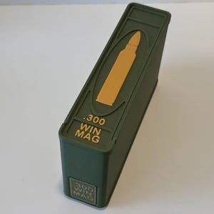 Puede incluir: Un contenedor rectangular verde con un gráfico de bala dorada y el texto ".300 WIN MAG". El contenedor está en ángulo, mostrando la parte superior y lateral, sobre un fondo blanco.