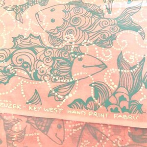 Peut inclure: Tissu rose avec un motif de poissons turquoise. Les poissons sont stylis&eacute;s avec des d&eacute;tails tourbillonnants et des lignes pointill&eacute;es blanches. Le texte "ZUZEK - KEY WEST HAND PRINT FABRIC" est imprim&eacute;.