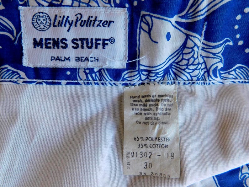 Peut inclure: Gros plan sur un tissu &agrave; motifs bleu et blanc avec une doublure blanche. Le tissu pr&eacute;sente un motif de poisson. Les &eacute;tiquettes indiquent "Lilly Pulitzer MENS STUFF PALM BEACH" et "65% POLYESTER 35% COTTON". La taille est de 76 cm.