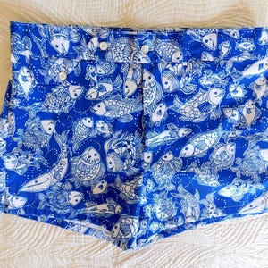 Peut inclure: Short bleu avec un motif de poissons blancs. Le short a une fermeture boutonn&eacute;e et est fait d'un tissu l&eacute;ger. Le motif de poissons comprend diverses esp&egrave;ces et tailles, cr&eacute;ant un look estival et ludique.