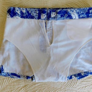 Peut inclure: Short de bain blanc avec un motif de poissons bleu et blanc sur les bords. Le short a une doublure blanche et une fermeture &eacute;clair &agrave; l'avant. Le tissu semble l&eacute;ger, adapt&eacute; &agrave; la natation. Mesures en centim&egrave;tres.