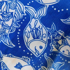 Peut inclure: Gros plan sur un tissu avec un motif de poissons bleu et blanc. Les poissons sont soulign&eacute;s en bleu sur un fond blanc, avec un design stylis&eacute;. Le mot "Lilly" est &eacute;crit &agrave; l'int&eacute;rieur d'un des poissons.