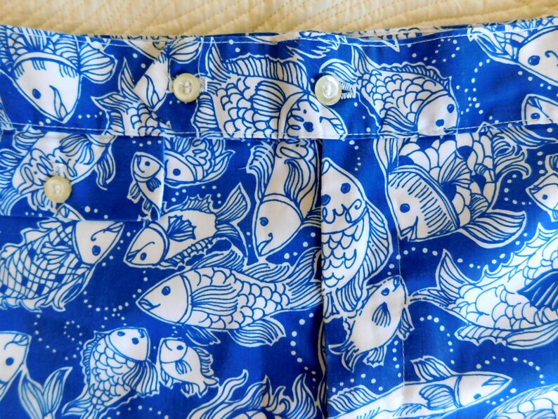 Peut inclure: Gros plan sur un tissu bleu avec un motif de poissons blancs. Le tissu pr&eacute;sente un motif r&eacute;p&eacute;t&eacute; de poissons stylis&eacute;s, avec des d&eacute;tails tels que des nageoires et des &eacute;cailles. Le tissu est fronc&eacute; et comporte des boutons visibles.