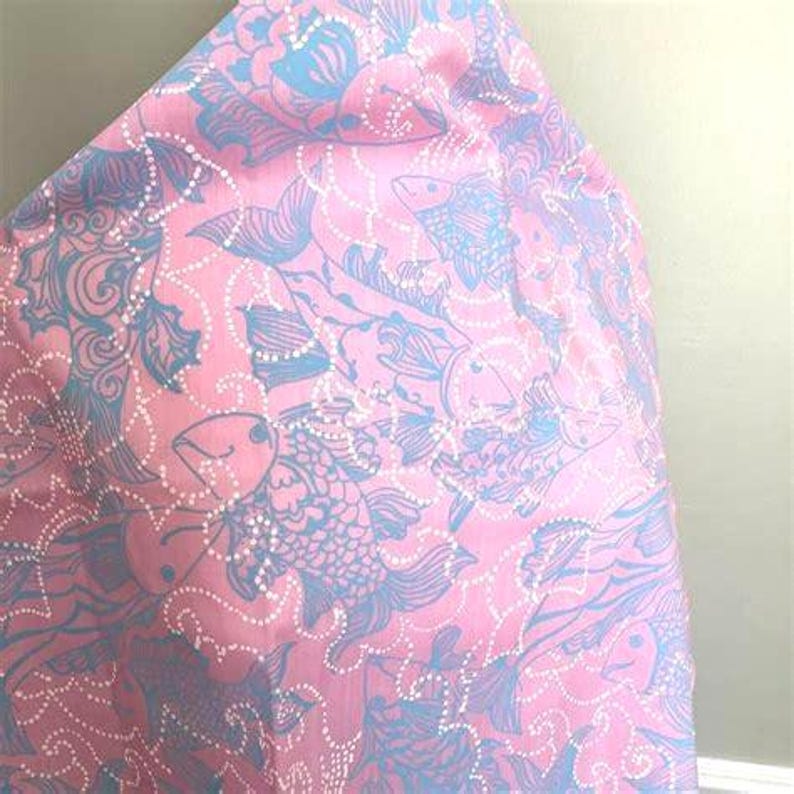 Peut inclure: Tissu rose avec un motif r&eacute;p&eacute;titif de poissons bleus et de motifs floraux. Les poissons sont d&eacute;taill&eacute;s avec des lignes pointill&eacute;es blanches, cr&eacute;ant un th&egrave;me aquatique fantaisiste. Le tissu est pli&eacute;, montrant la continuit&eacute; du motif.