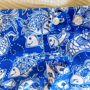 Peut inclure: Gros plan sur un tissu bleu et blanc avec un motif de poissons. Le tissu pr&eacute;sente un motif r&eacute;p&eacute;t&eacute; de poissons stylis&eacute;s en blanc sur un fond bleu vif. Deux boutons blancs sont visibles sur le tissu.