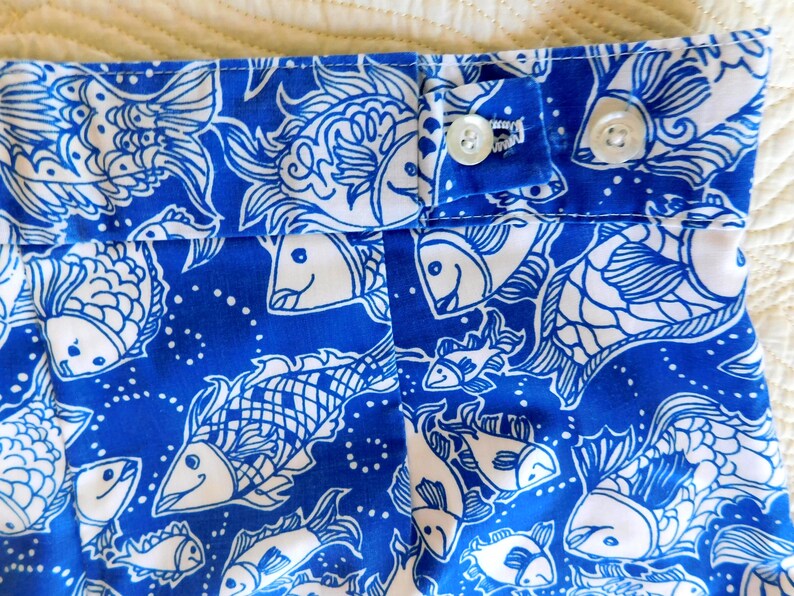 Peut inclure: Gros plan sur un tissu bleu et blanc avec un motif de poissons. Le tissu pr&eacute;sente un motif r&eacute;p&eacute;t&eacute; de poissons stylis&eacute;s en blanc sur un fond bleu vif. Deux boutons blancs sont visibles sur le tissu.