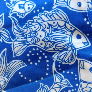 Peut inclure: Gros plan sur un tissu bleu avec un motif de poissons blancs. Les poissons sont stylis&eacute;s avec des nageoires et des &eacute;cailles d&eacute;taill&eacute;es, et le fond pr&eacute;sente de petits points blancs. Le motif se r&eacute;p&egrave;te sur le tissu.