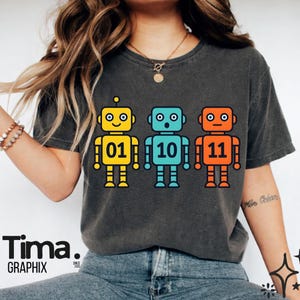 May include: Dark gray t-shirt featuring three cartoon robots in yellow, blue, and orange. Each robot has a number: 01, 10, and 11. The shirt has the text "Tima. GRAPHIX" at the bottom.