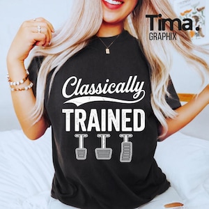 Pode incluir: T-shirt preto com o texto branco "Classically TRAINED", com ilustrações de três pedais de carro. A camisa tem gola redonda e é de estilo casual. O logotipo da marca "Tima. GRAPHIX" está no canto superior direito.