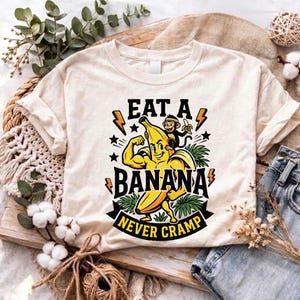 以下が含まれることがあります： クリーム色のTシャツで、腕を曲げて筋肉を見せている黄色いバナナのキャラクターと、その肩に乗った小さな猿のグラフィックが描かれています。テキストは「EAT A BANANA NEVER CRAMP」で、オレンジ色の稲妻と黒い星に囲まれています。