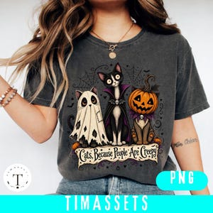 May include: A charcoal gray t-shirt featuring a graphic of three cats in Halloween costumes: one as a ghost, one as a vampire, and one with a pumpkin head. The text below reads, Cats, Because People Are Creepy, surrounded by spiderwebs.