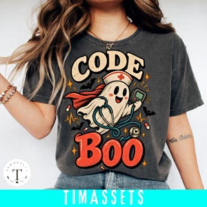 Puede incluir: Una camiseta gris carbón con el gráfico de un fantasma con cofia de enfermero y capa, sosteniendo un estetoscopio y un dispositivo médico. El texto CODE BOO aparece en letras grandes, rodeado de pequeños murciélagos, pastillas y líneas.