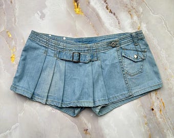 May include: A light blue denim mini skirt featuring a pleated design, a horizontal strap with a metal buckle at the waist, and a small flap pocket on the right side secured with a silver-toned metal snap button closure.