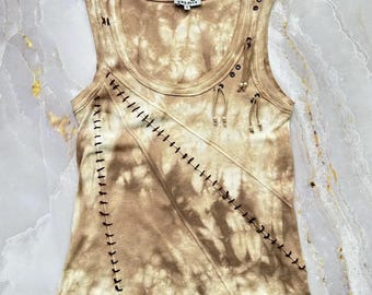 May include: A sleeveless tank top in a tan and brown tie-dye pattern featuring a scoop neckline and a label reading yasmin. The garment includes decorative dark brown whip-stitching along diagonal seams and small brown beads on thin fabric ties.