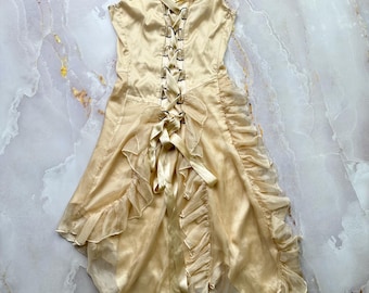 Puede incluir: Un vestido color crema con un corpiño estilo corsé y una falda con volantes. El vestido presenta una parte delantera con cordones, correas con ojales y un dobladillo asimétrico y fluido. La tela parece ser un material ligero y sedoso.