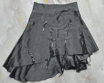 May include: A charcoal gray skirt with a high-low hemline and a shiny, iridescent finish. The skirt features a large front pocket detail adorned with thin, dangling ribbon ties attached to silver-toned metal hardware, creating a layered look.