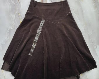 May include: A dark brown midi skirt featuring a high-waisted design with belt loops. The skirt has a diagonal seam across the front accented with small silver studs. A decorative vertical strap with two silver buckles and grommets hangs down.