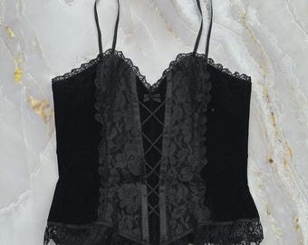 Puede incluir: Un top tipo corsé de terciopelo negro con tirantes finos, un panel central con cordones y un pequeño lazo, y un intrincado borde de encaje negro a lo largo del escote y el dobladillo. El corpiño tiene un diseño estructurado y un dobladillo puntiagudo.