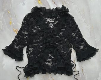 May include: A black lace top featuring a V-neckline, three-quarter length sleeves, and ruffled trim along the collar, front opening, and sleeve cuffs. The garment includes thin side ties and a sheer, intricate floral pattern throughout the fabric.