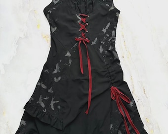 Puede incluir: Un vestido negro sin mangas con un patrón de aves en gris oscuro, escote cuadrado y un corpiño central tipo corsé con cordones rojos. La falda incluye un panel lateral fruncido con cintas rojas y un dobladillo con volantes.
