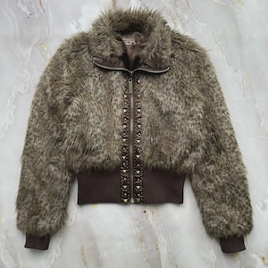 Puede incluir: Chaqueta de piel sint&eacute;tica marr&oacute;n con cremallera y detalles de tachuelas. La chaqueta tiene un dobladillo y pu&ntilde;os acanalados, y un cuello alto. La piel sint&eacute;tica es una mezcla de tonos marrones y grises, creando un aspecto texturizado. La chaqueta es de estilo corto.