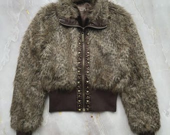 May include: A brown faux fur jacket with a zip-up front and studded detailing. The jacket has a ribbed hem and cuffs, and a high collar. The faux fur is a mix of brown and gray tones, creating a textured look. The jacket is a cropped style.