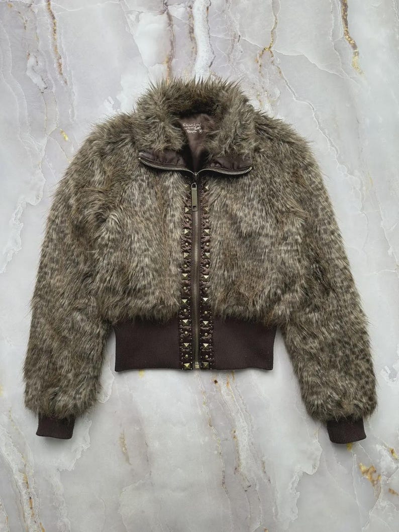 Puede incluir: Chaqueta de piel sint&eacute;tica marr&oacute;n con cremallera y detalles de tachuelas. La chaqueta tiene un dobladillo y pu&ntilde;os acanalados, y un cuello alto. La piel sint&eacute;tica es una mezcla de tonos marrones y grises, creando un aspecto texturizado. La chaqueta es de estilo corto.