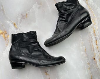 May include: A pair of black leather ankle boots featuring a ruched design on the upper shaft, a rounded toe, and a low block heel. The boots have visible stitching along the sides and a smooth finish, showcasing a classic silhouette with a side zipper.