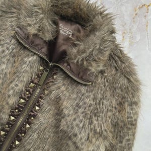 Puede incluir: Un chaleco de piel sint&eacute;tica en tonos marr&oacute;n y gris, con cuello alto y cierre de cremallera. El chaleco est&aacute; adornado con tachuelas doradas a lo largo de la cremallera, a&ntilde;adiendo un toque de estilo atrevido. El forro es de color marr&oacute;n oscuro.
