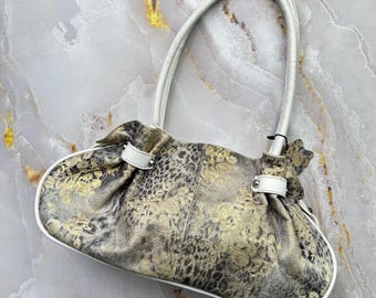 May include: A handbag featuring a grey and black animal print pattern with gold metallic accents. The bag has a curved silhouette, white leather trim, and two white leather shoulder straps attached with silver-toned metal hardware.