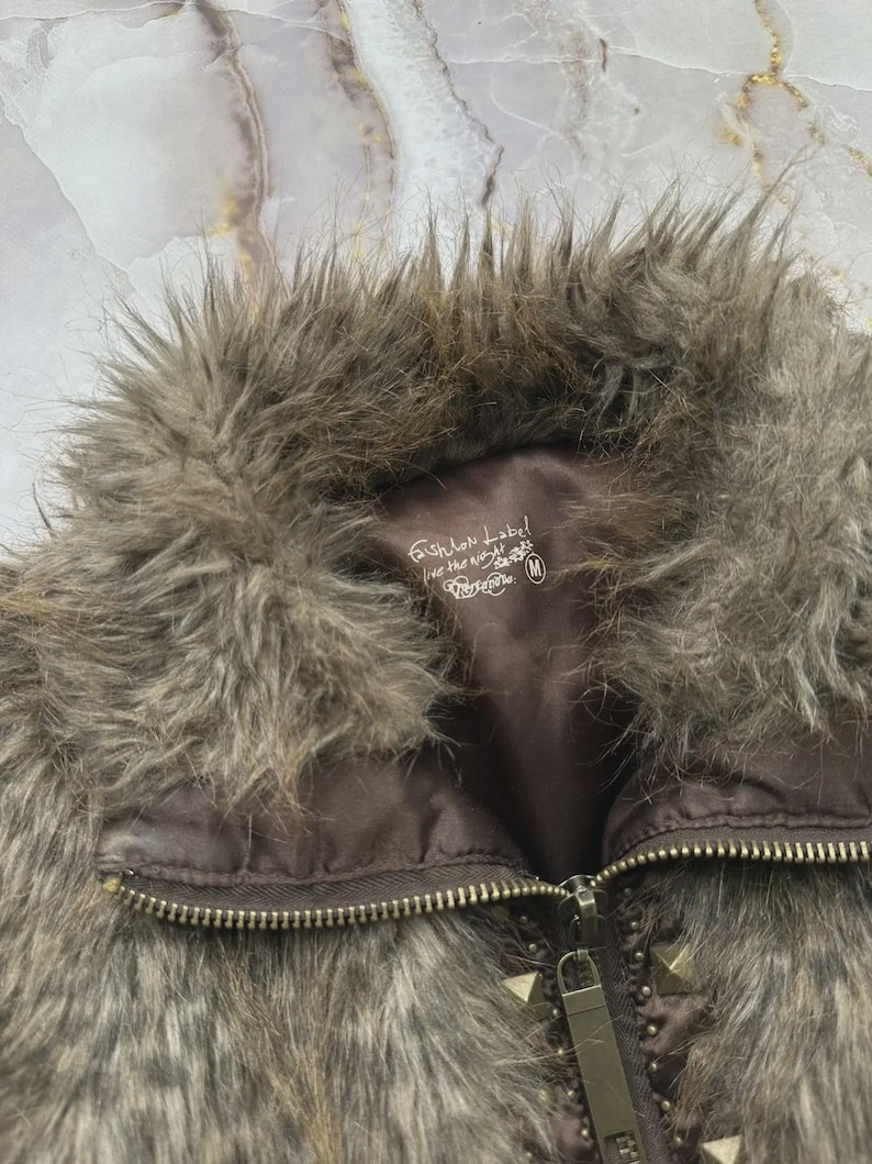 Puede incluir: Primer plano de un chaleco de piel sint&eacute;tica marr&oacute;n con cierre de cremallera y detalles de tachuelas. El chaleco tiene un cuello esponjoso y un forro marr&oacute;n. La etiqueta dice "Fashion Label, live the night".