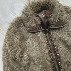 Puede incluir: Chaqueta de piel sint&eacute;tica marr&oacute;n con cierre de cremallera y detalles de tachuelas a lo largo de la cremallera. La chaqueta tiene un cuello alto y un aspecto suave y texturizado. El forro es de color marr&oacute;n oscuro.