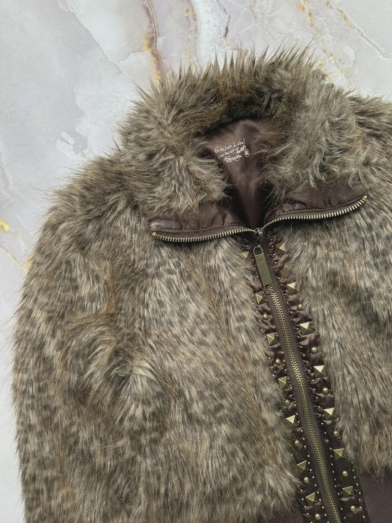 Puede incluir: Chaqueta de piel sint&eacute;tica marr&oacute;n con cierre de cremallera y detalles de tachuelas a lo largo de la cremallera. La chaqueta tiene un cuello alto y un aspecto suave y texturizado. El forro es de color marr&oacute;n oscuro.