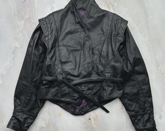 May include: A black leather jacket featuring a high collar, structured shoulders, and a wrap-style front closure secured with a thin leather belt. The jacket has long sleeves with cuffs and a visible purple lining visible at the neckline and hem.