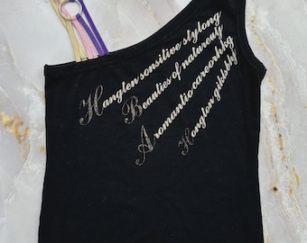 Puede incluir: Una camiseta de tirantes negra de un solo hombro con tres tirantes finos en amarillo, rosa y morado conectados por un anillo de metal plateado. El frente muestra texto plateado brillante que dice Hangten sonsitive stylong Beauties of natureuy A romantio careorhkg Hongton giklslhj.