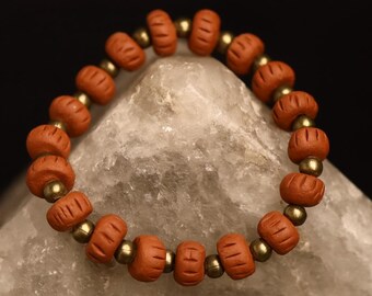 May include: A terracotta-colored beaded bracelet with gold-colored spacers. The bracelet is resting on a light gray, geometric-shaped surface against a dark background. The beads have a carved design, and the bracelet is circular.
