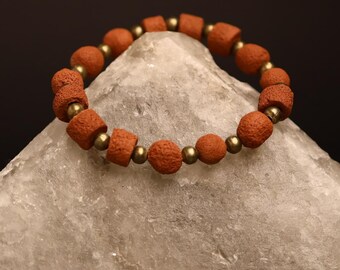 May include: A beaded bracelet with reddish-brown beads and gold-colored spacers rests on a light gray, square-shaped stone. The bracelet is circular, and the beads are evenly spaced. The background is a solid black.