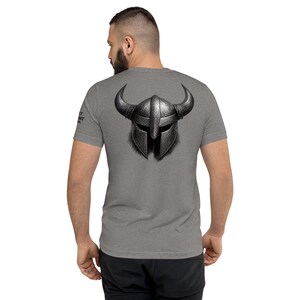 Könnte beinhalten: Graues T-Shirt mit einer detaillierten Illustration eines Wikingerhelms mit großen Hörnern auf der Rückseite. Der Helm ist in Grau- und Schwarztönen gehalten und hat ein strukturiertes Aussehen. Das T-Shirt hat kurze Ärmel und einen Rundhalsausschnitt.