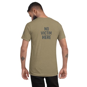 Könnte beinhalten: Olivgrünes T-Shirt mit dem Text "NO VICTIM HERE" in Dunkelgrau auf der Rückseite aufgedruckt. Das Shirt ist ein kurzärmeliges Rundhals-Modell. Das Model trägt eine schwarze Hose.