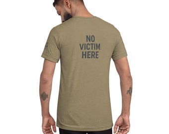May include: Olive green t-shirt with the text "NO VICTIM HERE" printed in dark gray on the back. The shirt is a short-sleeved crew neck style. The model is wearing black pants.