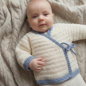 May include: A cream-colored, hand-knitted baby cardigan with light blue trim and a tie closure. The sweater has long sleeves and is made of a soft, textured yarn. The baby is lying on a beige cable-knit blanket.