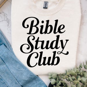 Op de afbeelding: Crèmekleurige sweatshirt met de tekst "Bible Study Club" in een vet zwart lettertype. De sweatshirt is opgevouwen, met een spijkerbroek en een stapel boeken op de achtergrond. Een takje groen is ook aanwezig.