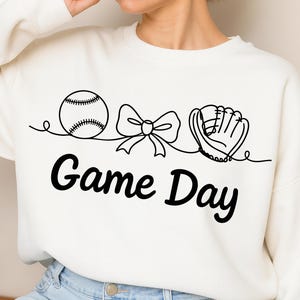 Op de afbeelding: Crèmekleurige sweatshirt met een zwarte lijntekening van een honkbal, een strik en een honkbalhandschoen. Onder de tekening staat de tekst "Game Day" in een vetgedrukte, zwarte letter. De sweatshirt is een casual, sportieve stijl.