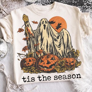 May include: A cream-colored t-shirt featuring a graphic of a ghost holding a knife, surrounded by orange pumpkins and falling autumn leaves against an orange moon. The text 'tis the season' is printed in black lettering at the bottom of the design.