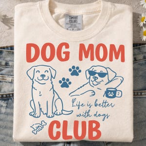 Dog Mom Club t-shirt mockup featuring cute dog illustrations, paw prints, and “Life is better with dogs” quote. Displayed on a folded cream shirt, perfect for dog moms, pet lovers, and gift ideas. Ideal for sublimation and print-on-demand.