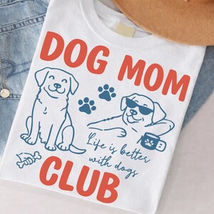 Dog Mom Club t-shirt mockup styled with denim and accessories, featuring cute dog illustrations and paw prints. Includes “Life is better with dogs” typography. Perfect for pet lovers, dog moms, and gift items.